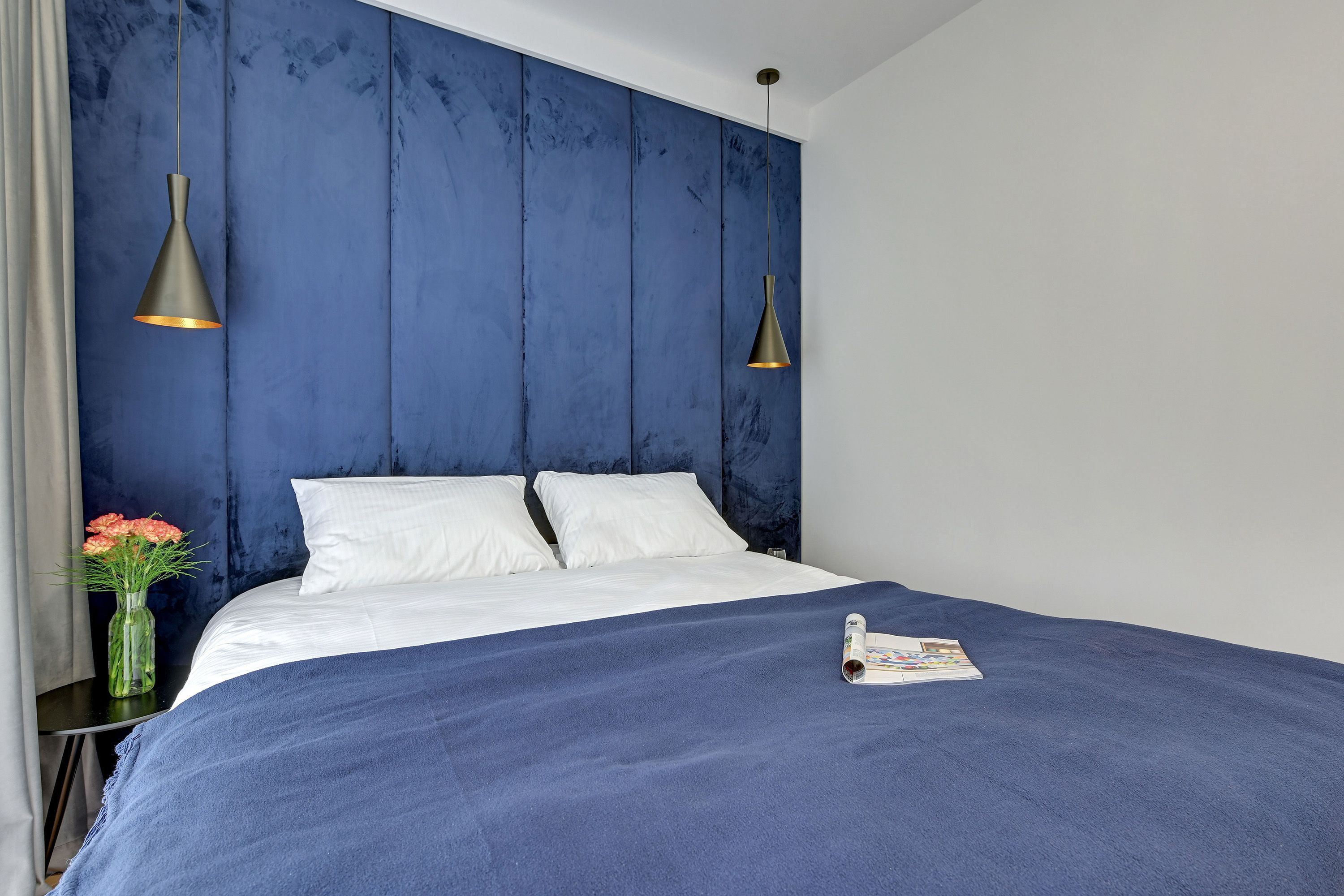 Una cama bien hecha con sábanas blancas, una colcha azul y dos lámparas colgantes.