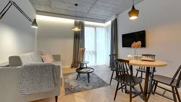 A modern living room with a grey sofa, a round wooden dining table with chairs, a black floor lamp, and a flat-screen TV mounted on the wall.