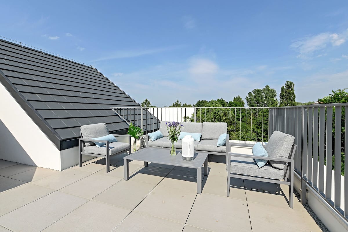 A rooftop terrace with a modern design, featuring a grey sofa, two armchairs, a coffee table, and a white vase with flowers.