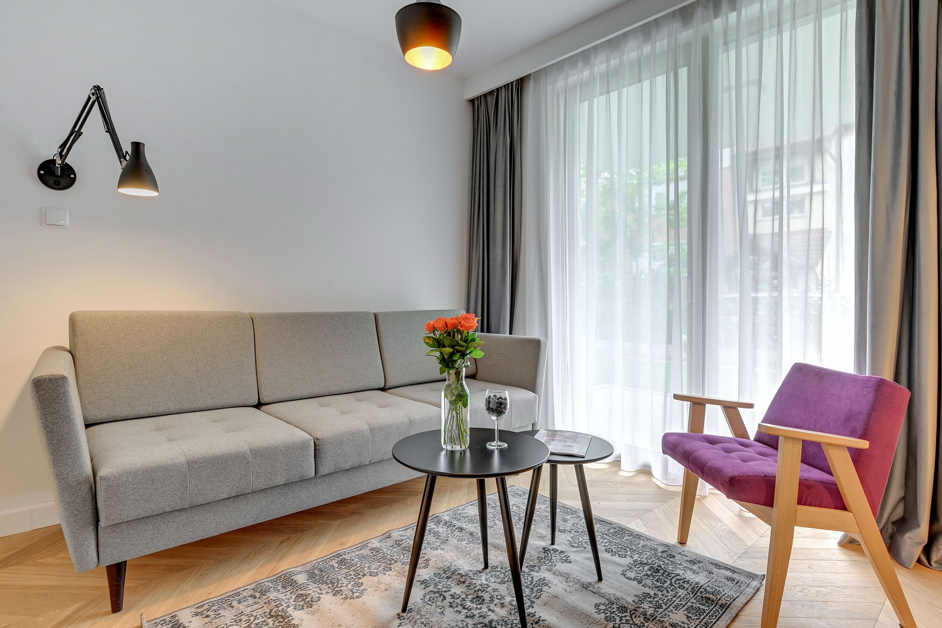 A modern living room with a grey sofa, a purple armchair, a small round table with a vase of flowers, and a floor lamp.