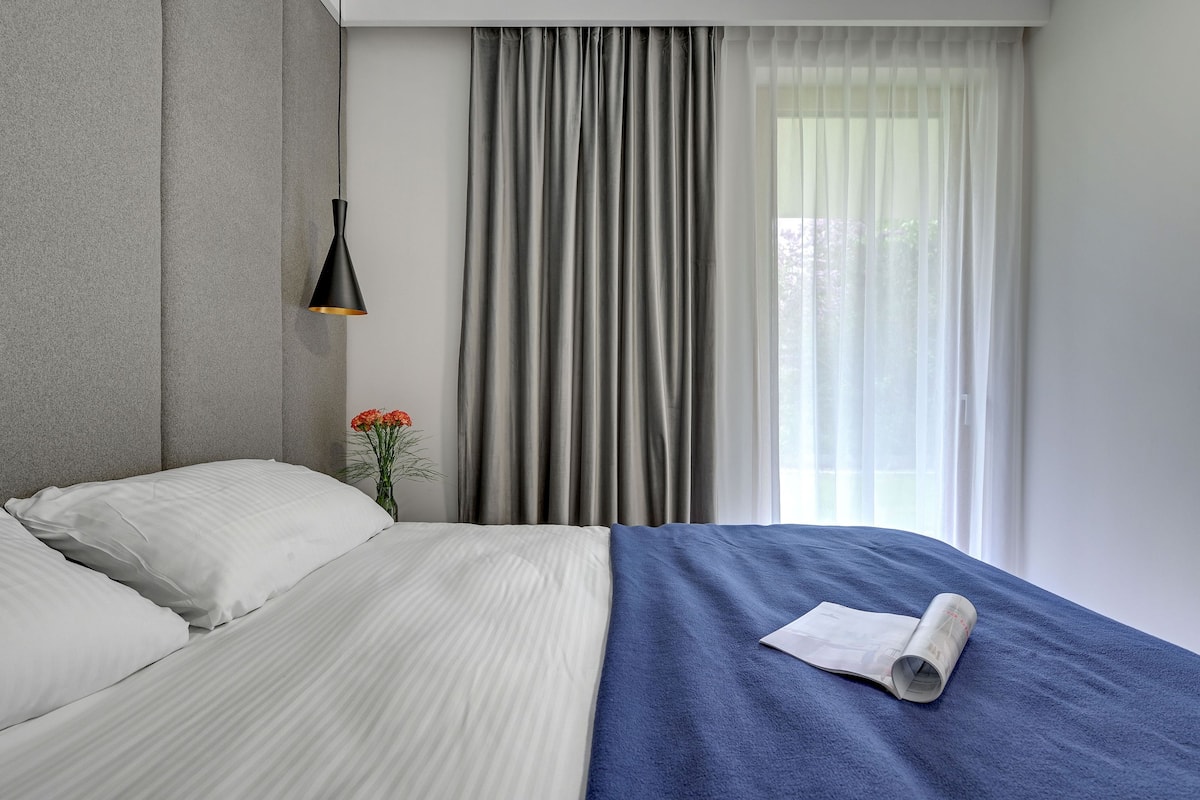 A neatly made bed with white and blue bedding, a black pendant light, grey curtains, and a window with sheer white curtains.