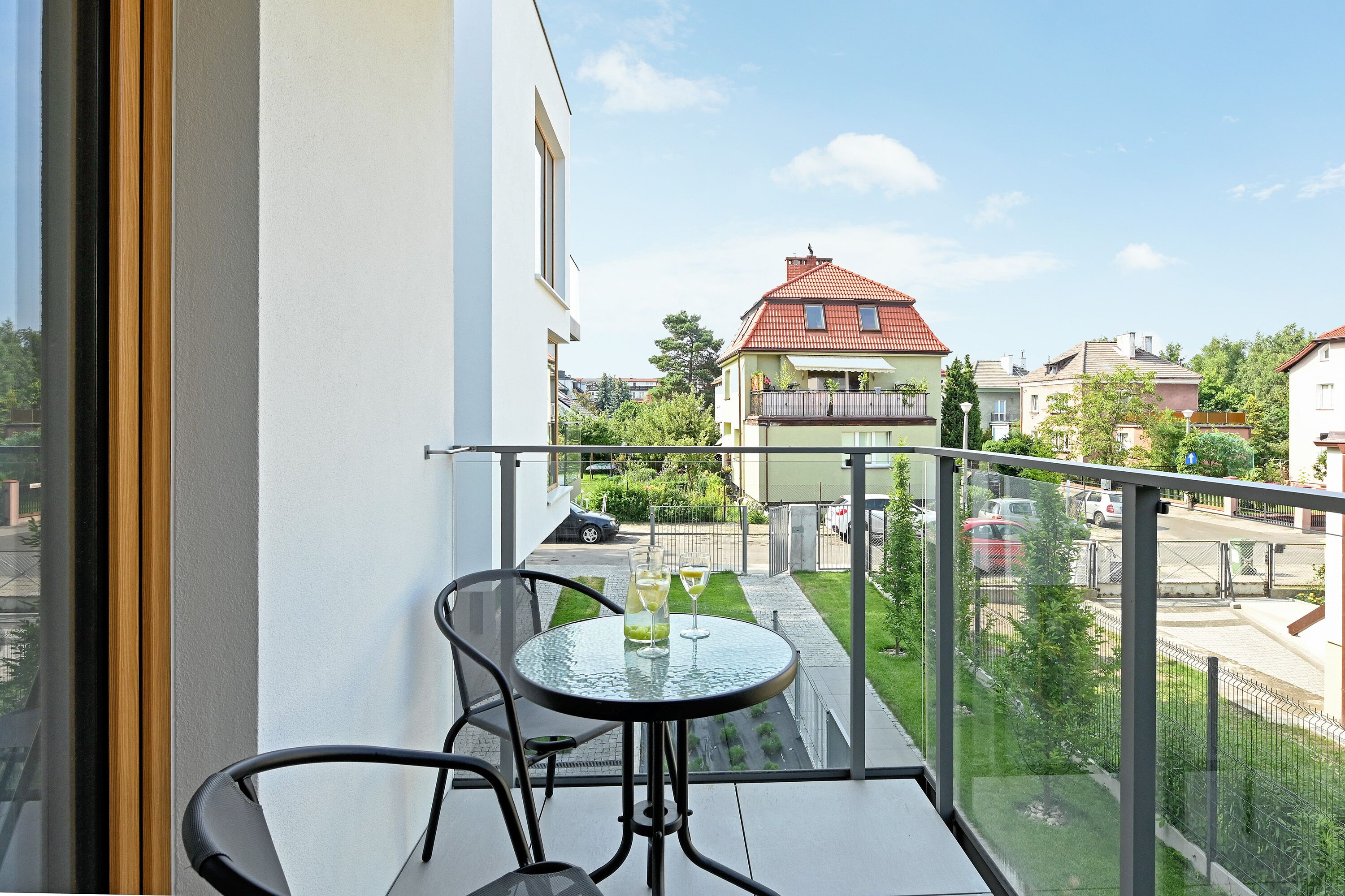 A balcony with a table and chairs, overlooking a street with houses and a clear sky.