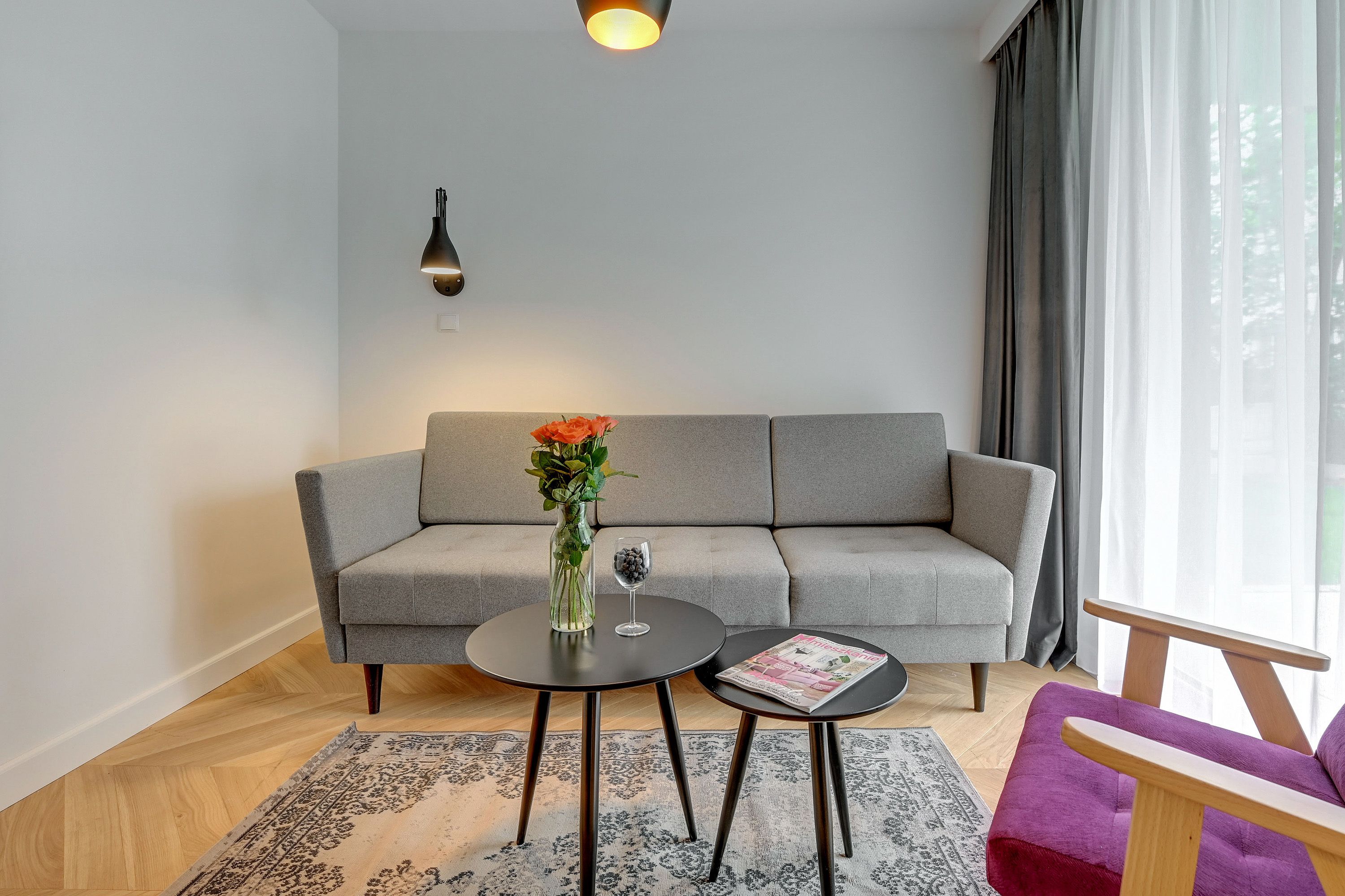 Een moderne woonkamer met een grijze bank, twee zwarte tafels, een paarse fauteuil en een groot raam met gordijnen.