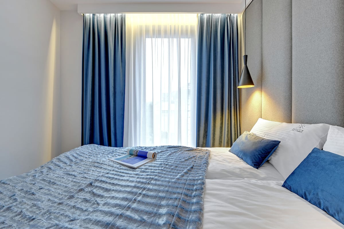 A modern hotel room with a neatly made bed, a large window with curtains, and a wall-mounted lamp.