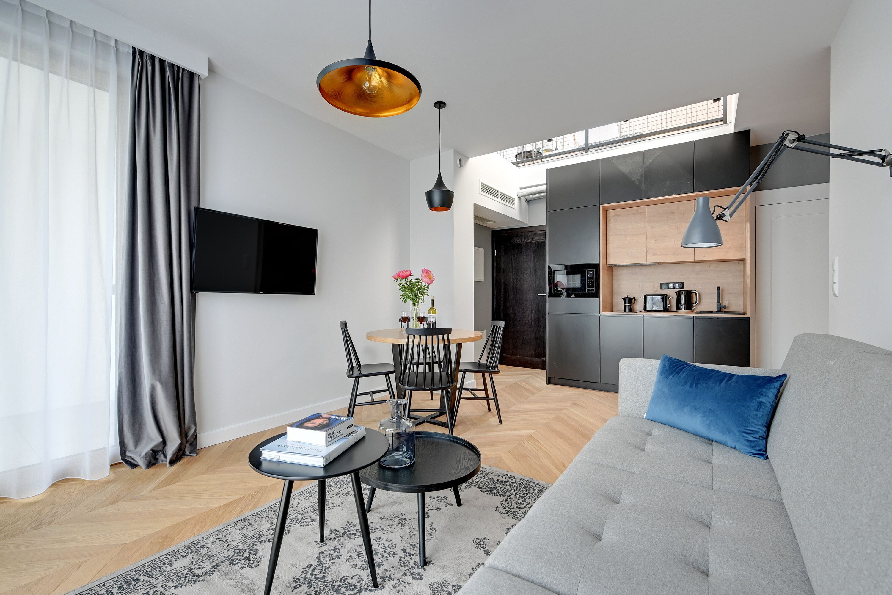 A modern living room with a grey sofa, a coffee table, a TV mounted on the wall, and a dining area with a table and chairs.