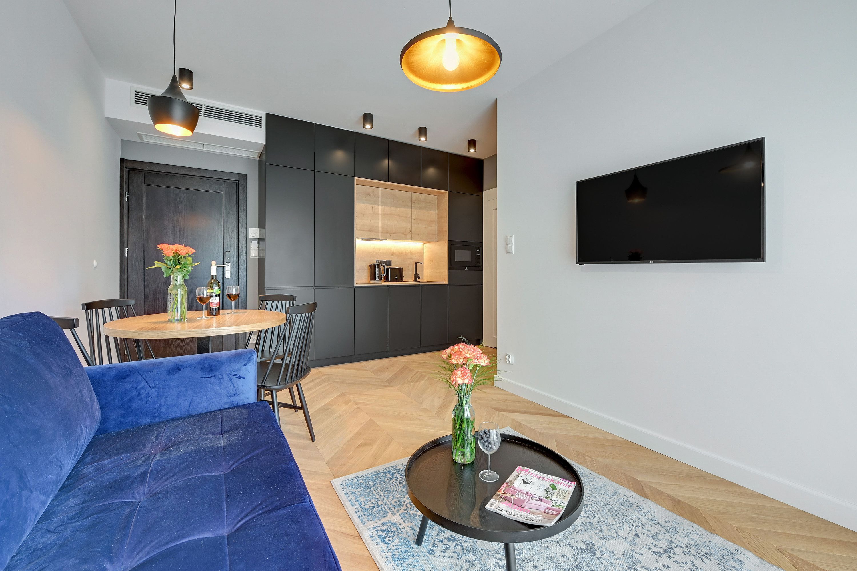 A modern living room with a blue sofa, a round coffee table with a magazine, a vase with flowers, and a TV mounted on the wall.