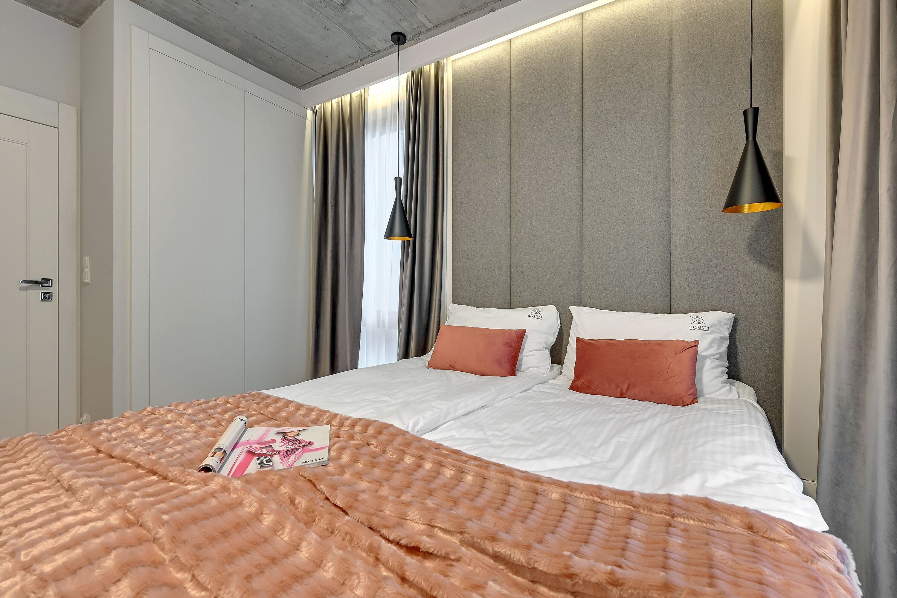 A modern hotel room with a large bed, a fluffy orange blanket, and a magazine on the bed. There is a grey headboard, a black pendant light, and a window with curtains.