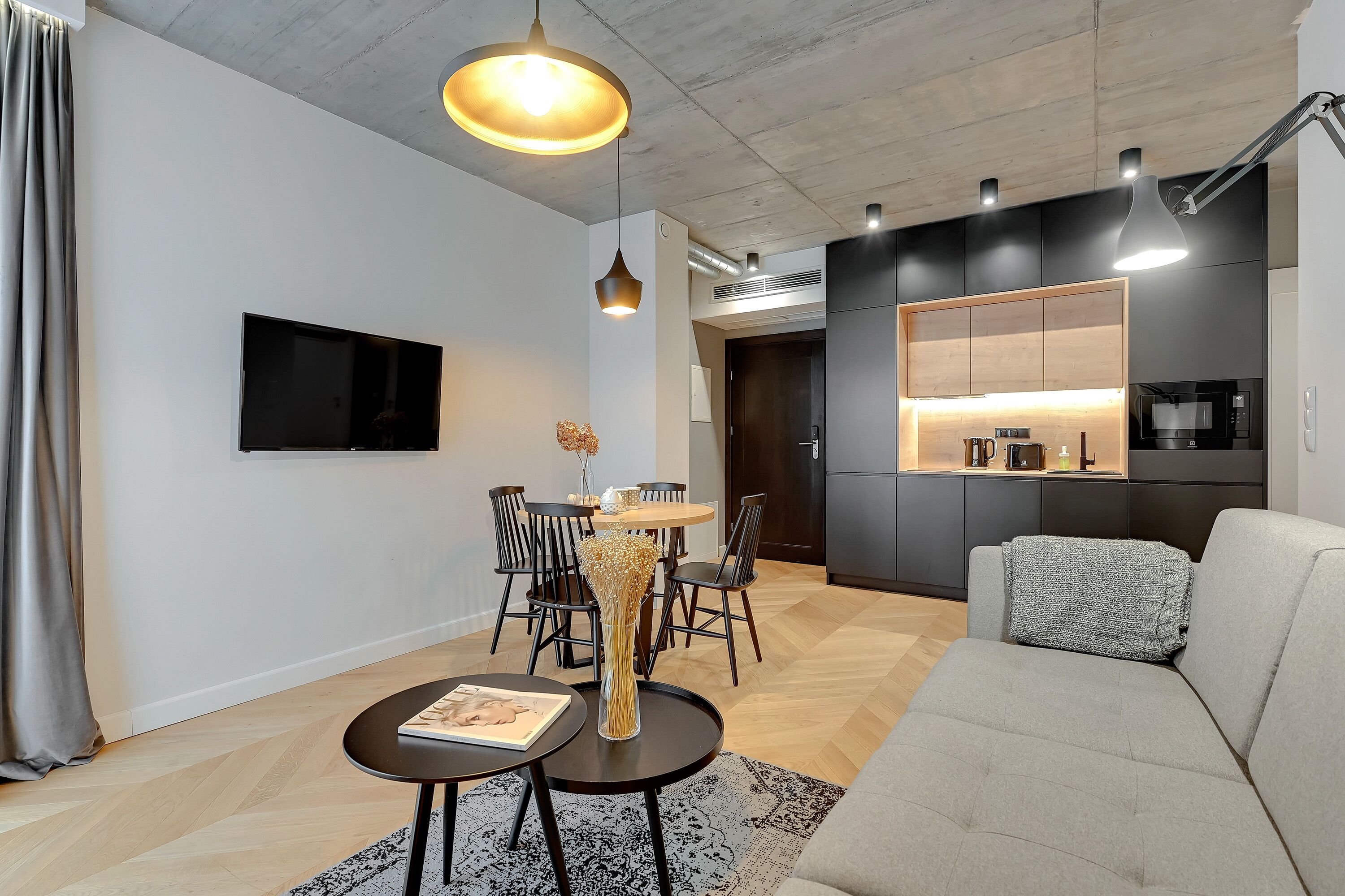Una sala de estar moderna con un sofá, una mesa de comedor con sillas, un televisor en la pared y una zona de cocina con microondas y armarios.