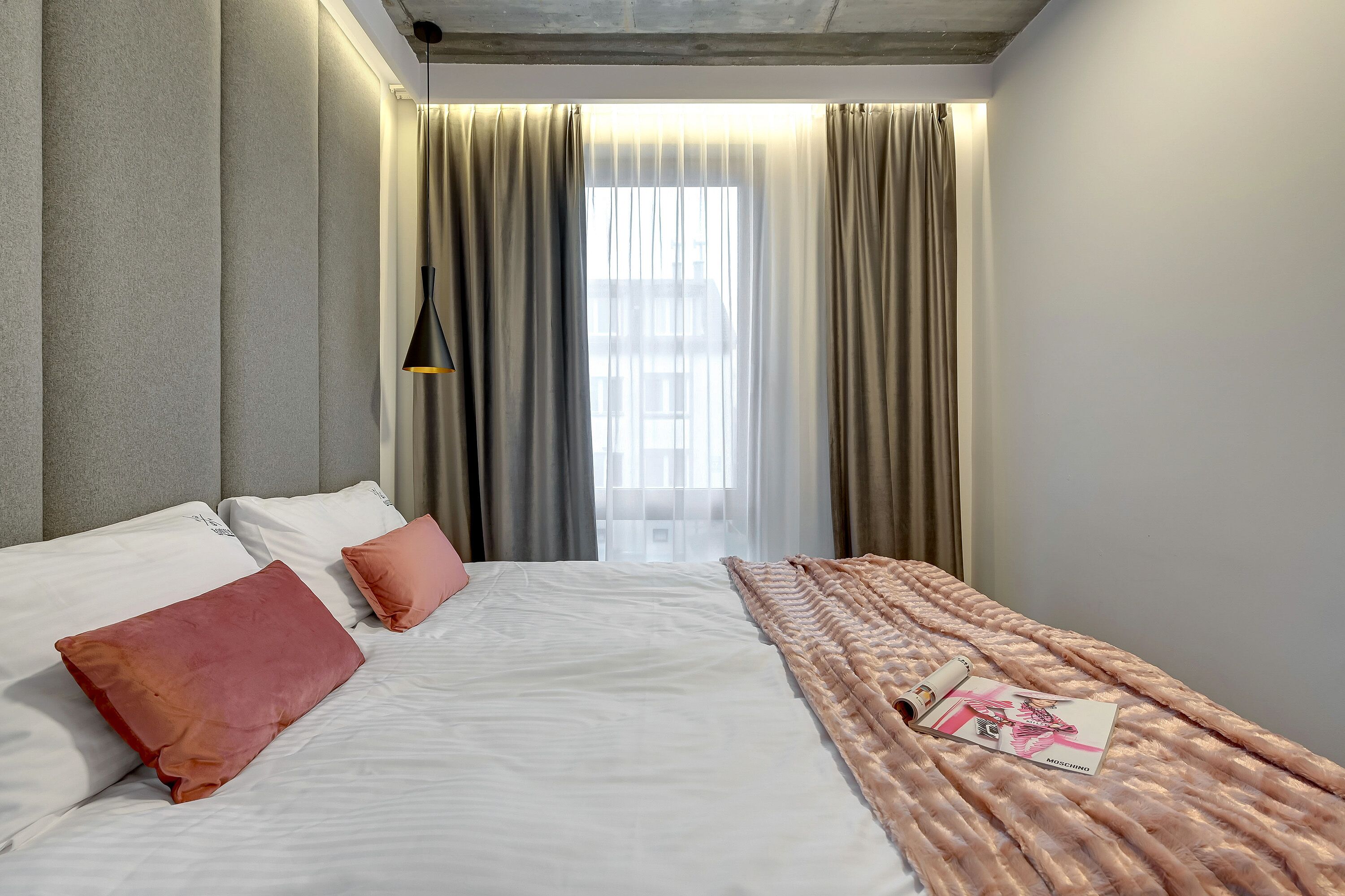 Un lit bien fait, avec des oreillers blancs et roses, un magazine posé sur le lit et une fenêtre avec des rideaux.
