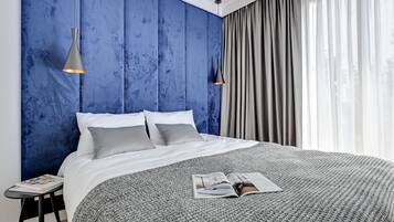 Een moderne slaapkamer met een groot bed, twee bedlampen, een tijdschrift op het bed en uitzicht op het landschap door dunne gordijnen.