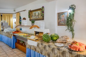 A buffet setup with various food items, a menu board, and decorative elements like a painting and a plant. - Palm Bay (Agios Nikolaos)