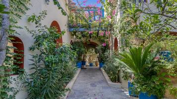 A narrow stone pathway flanked by lush greenery and potted plants, leading to a building with arched windows and a blue structure above.