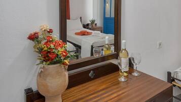 A room with a wooden dresser, a vase of flowers, a bottle of wine, and a mirror reflecting a bed and a blue door.
