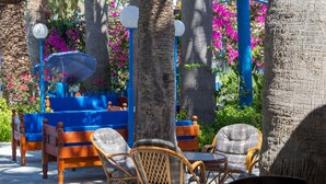 An outdoor seating area with wicker chairs and a blue sofa, surrounded by trees and vibrant pink flowers. - Palm Bay (Agios Nikolaos)