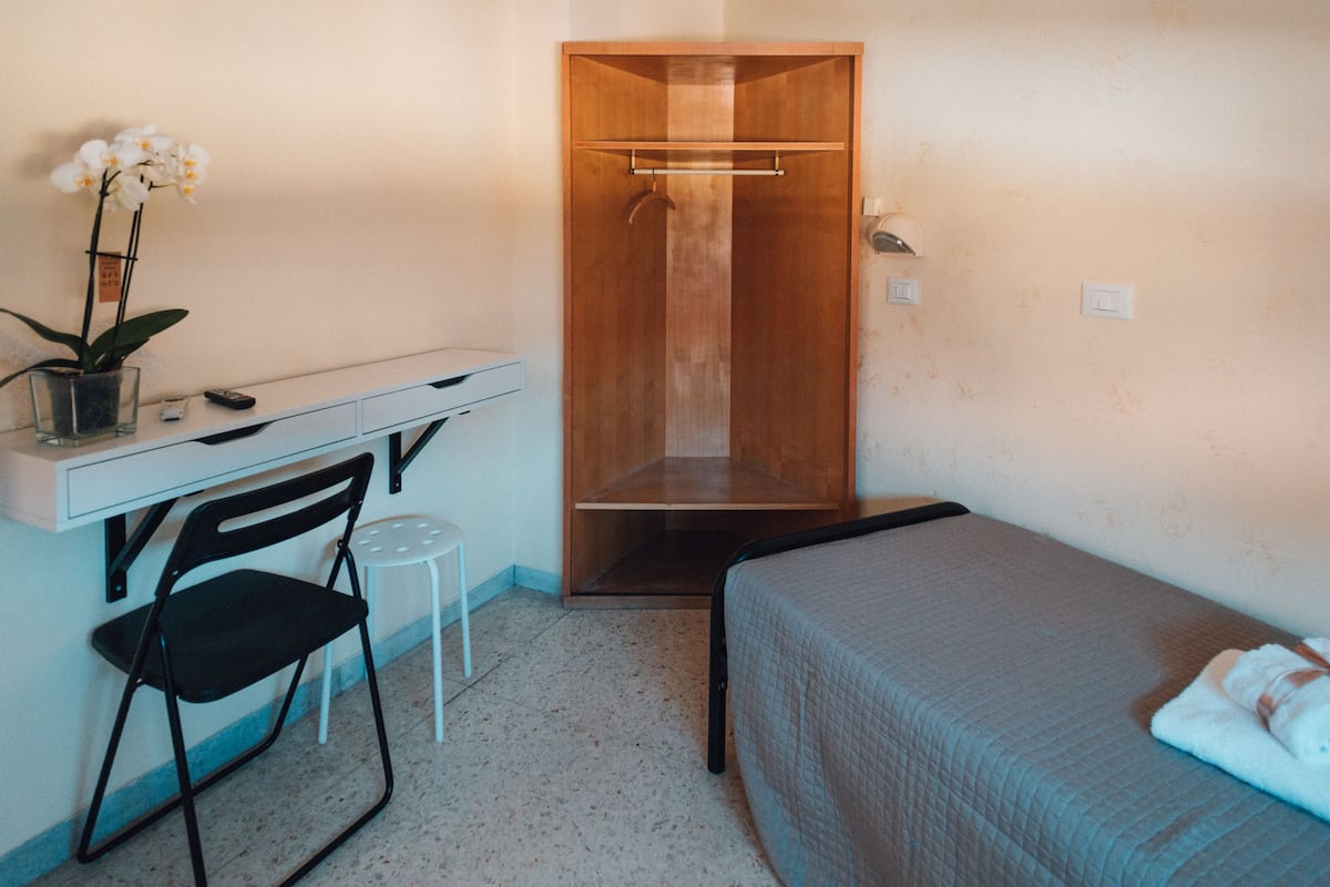 A small, compact room with a single bed, a desk with a chair, a wooden wardrobe, and a small potted plant.