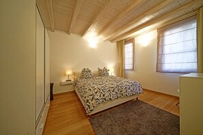 A bedroom with a bed, two bedside lamps, a window with curtains, and a wooden ceiling.