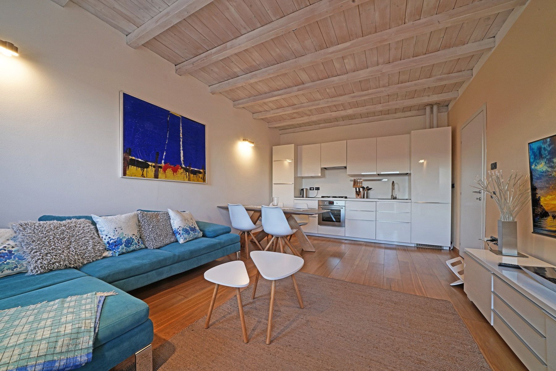 A modern living room with a blue sofa, a dining area with white chairs, a kitchen with white cabinets, and a wooden ceiling.