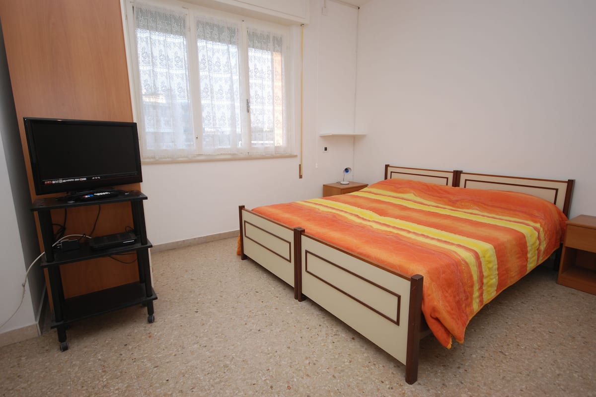 A bedroom with a bed, a television on a stand, a nightstand, and a window with curtains.