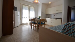 A compact kitchen with white cabinets, a dining table with chairs, and a small window with curtains. - Villa Gaby (Lignano Sabbiadoro)