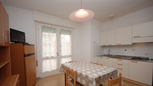 A compact kitchen with a dining table, chairs, a refrigerator, and a television on a cabinet. - Villa Gaby (Lignano Sabbiadoro)