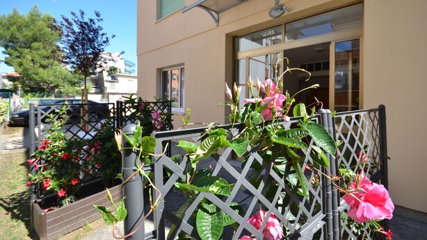 A building with a beige facade, a window with a metal frame, and a door. There is a metal fence with a lattice design, and a tree is visible in the background. - Villa Gaby (Lignano Sabbiadoro)