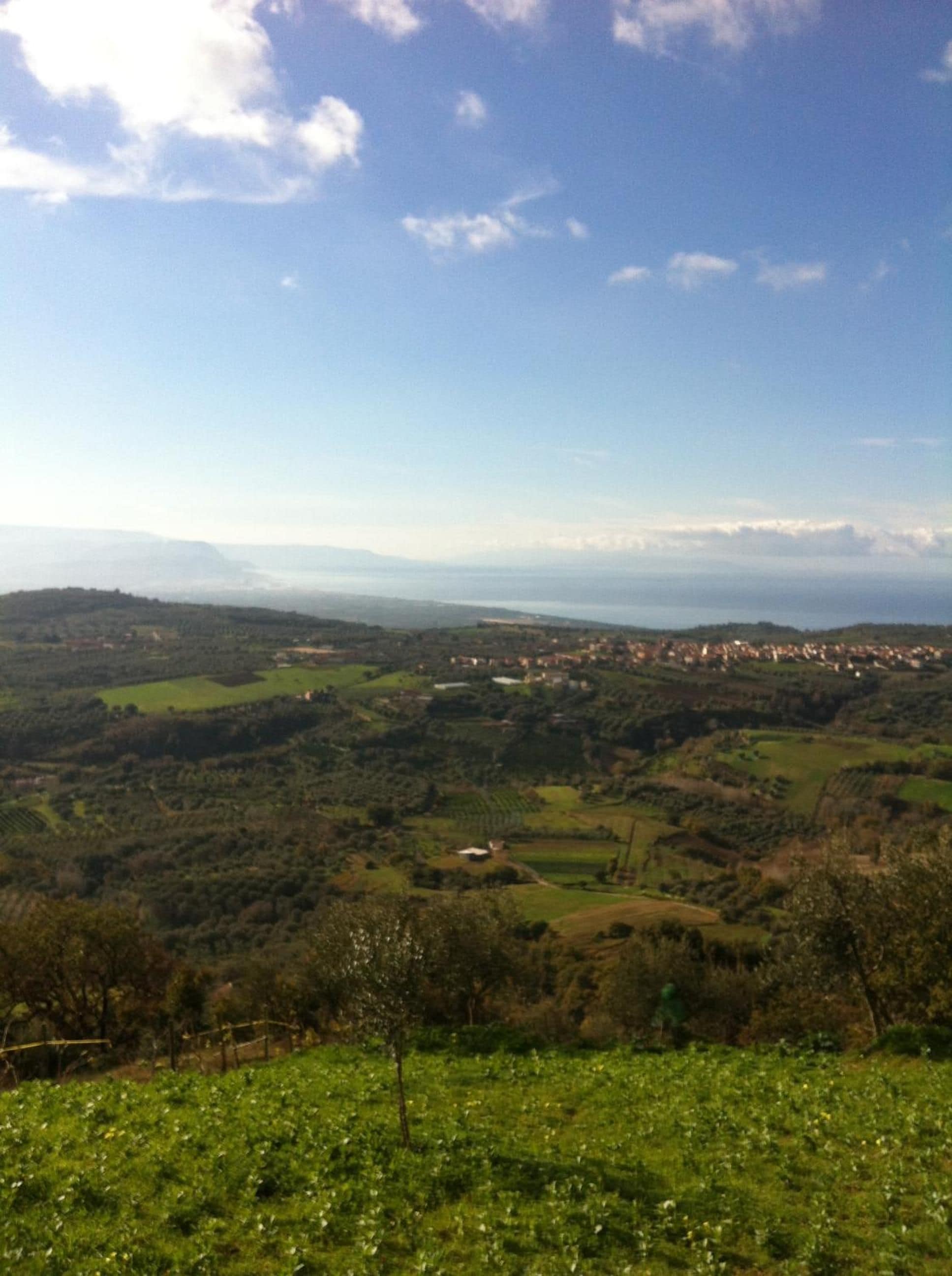 Vista panorámica de un paisaje montañoso con un pueblo a lo lejos, rodeado de vegetación y campos.