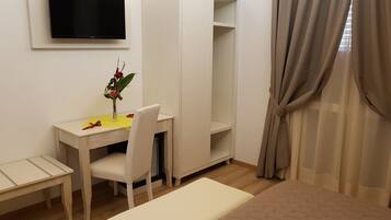 A small, neatly arranged room with a white desk, chair, and bench, a wardrobe, a television mounted on the wall, and a window with curtains.