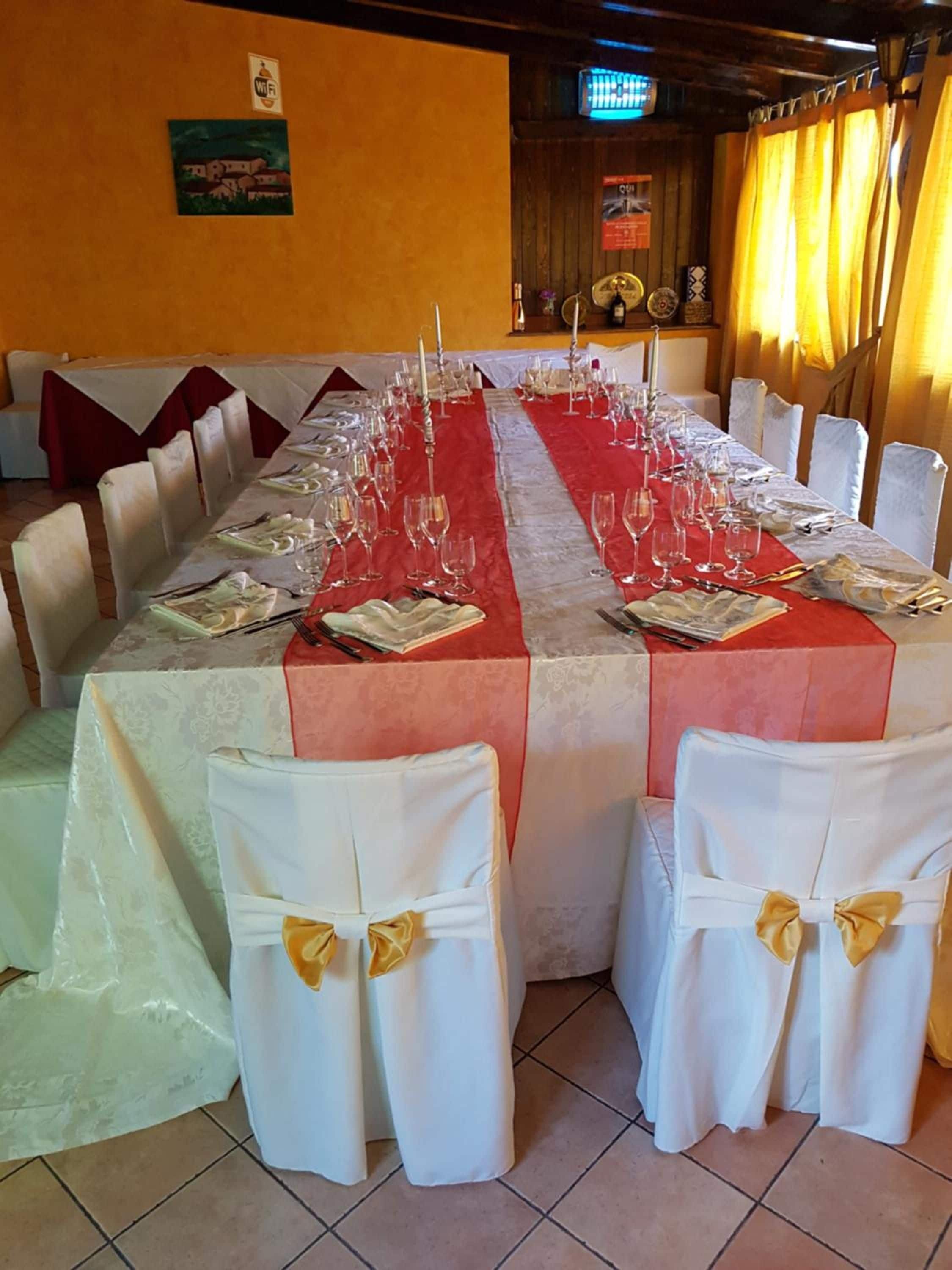 A long dining table set with multiple chairs, each with a white chair cover and a gold bow. The table is covered with a red tablecloth and has wine glasses and cutlery arranged.