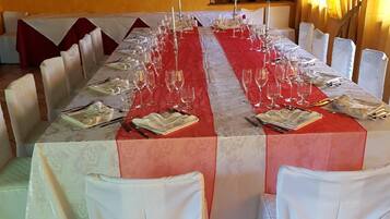 A long dining table set with multiple chairs, each with a white chair cover and a gold bow. The table is covered with a red tablecloth and has wine glasses and cutlery arranged.
