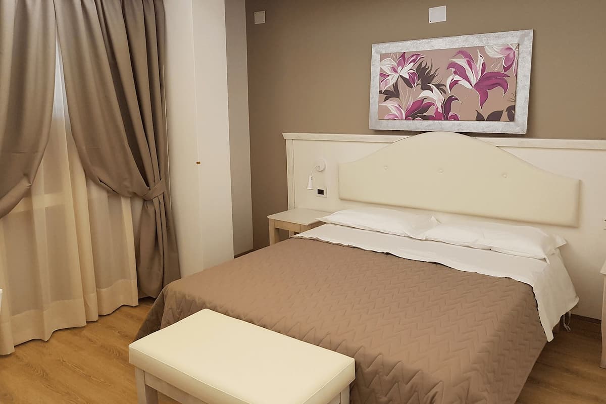 A bedroom with a bed, a bedside table, a nightstand, a bench, a wall-mounted lamp, and a painting.