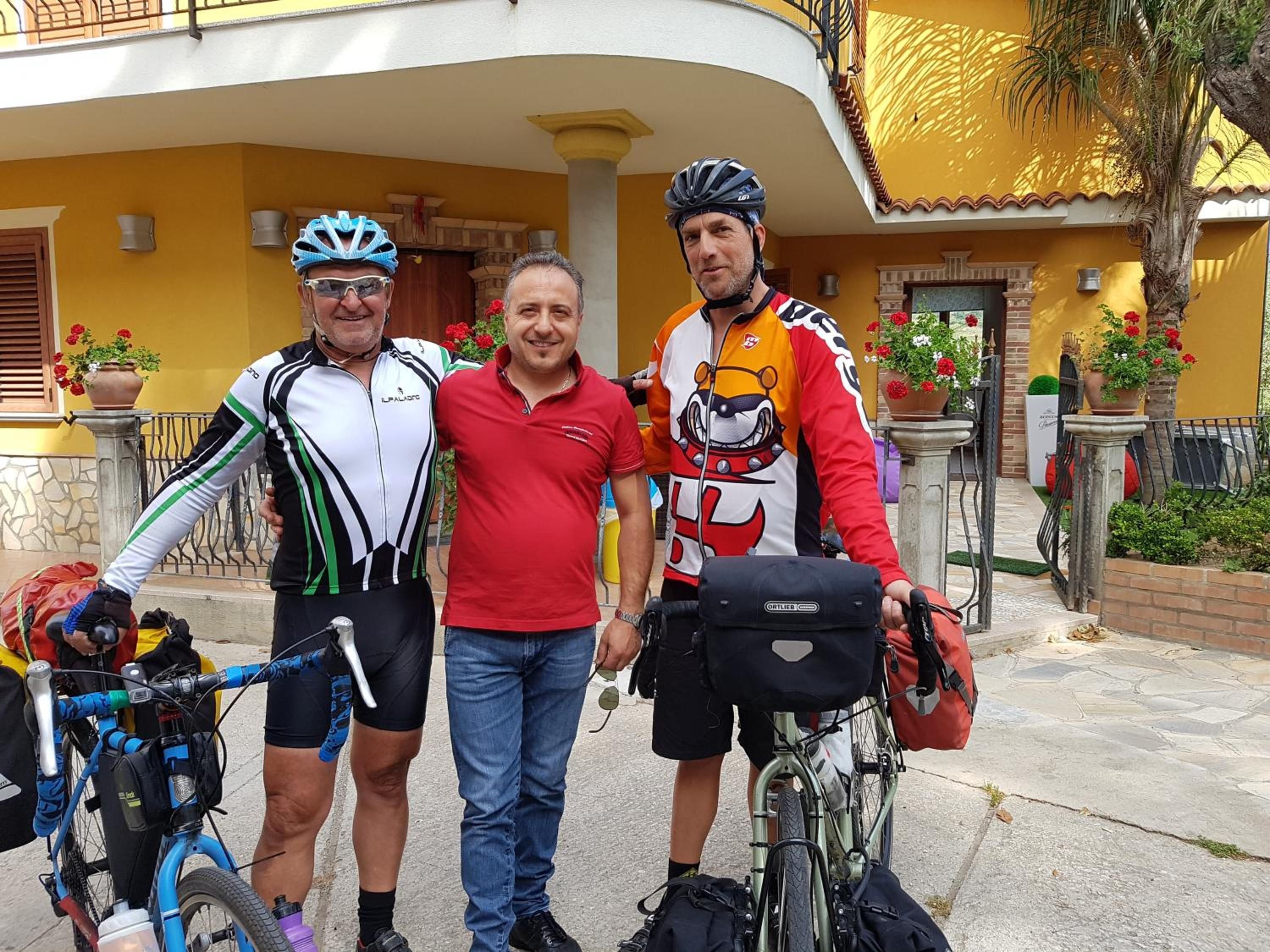 Tres personas de pie frente a un edificio amarillo con un balcón, una planta en maceta y una bicicleta.