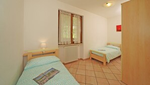 A small, well-lit room with two single beds, a window with sheer curtains, a radiator, and a small table with a lamp.