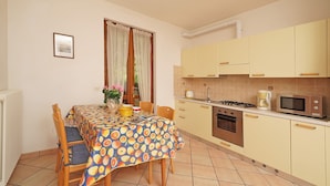 A dining area with a table covered in a fruit pattern tablecloth, wooden chairs, and a vase with flowers.