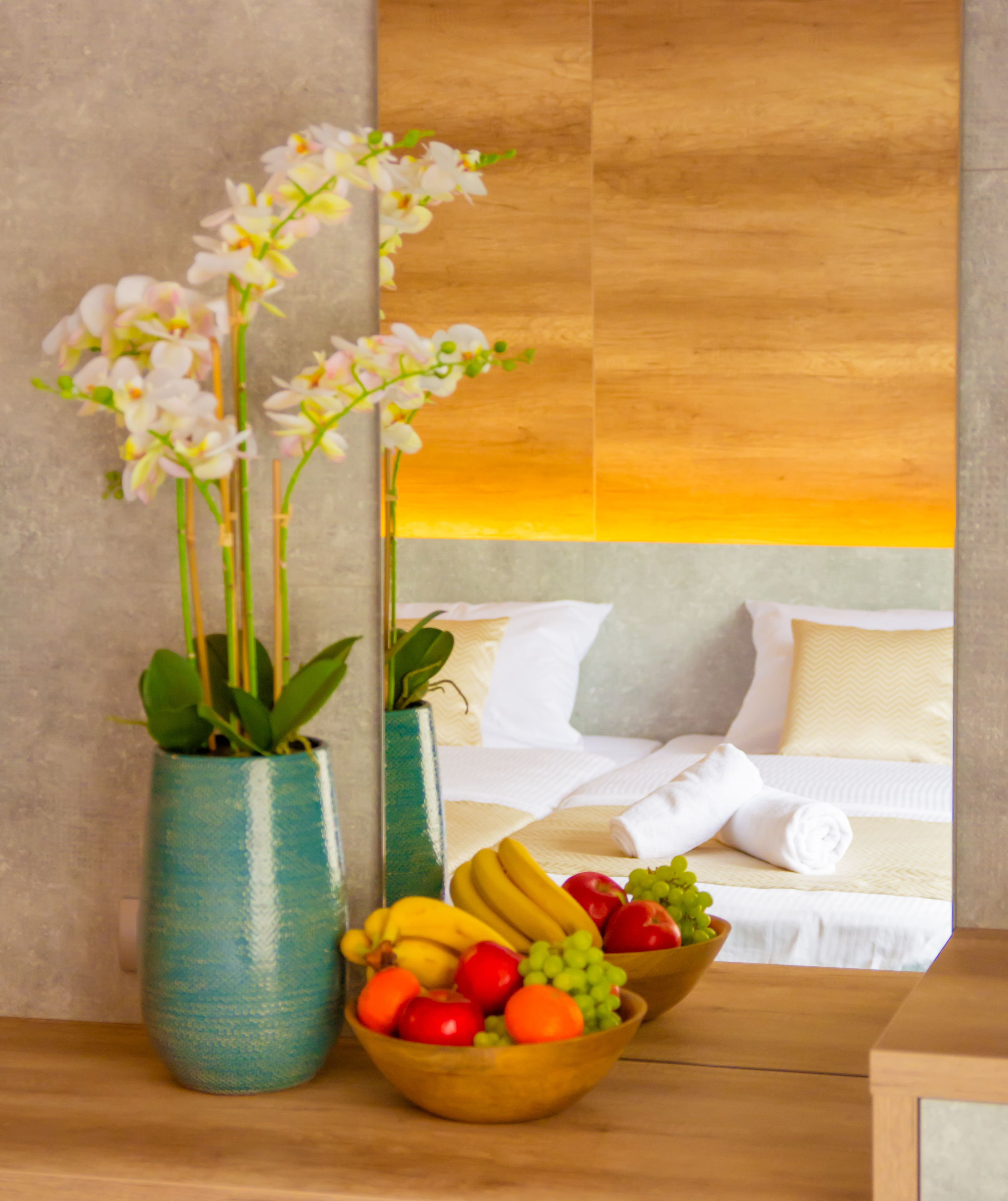 Une chambre d’hôtel avec un lit, des oreillers, un vase de fleurs et un panier de fruits.