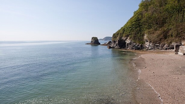 Ein Strand mit klarem Wasser, einem Felsen und einer bewaldeten Klippe.