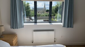 Ein Schlafzimmer mit großem Fenster, Heizkörper, Nachttisch aus Holz und einem Bett.
