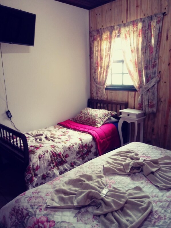 Une petite chambre avec deux lits, une tête de lit en bois, une table de chevet et une fenêtre ornée de rideaux à motifs floraux.