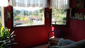 Una habitación con pared roja, ventana con cortinas blancas y vista a un pueblo, además de una planta.