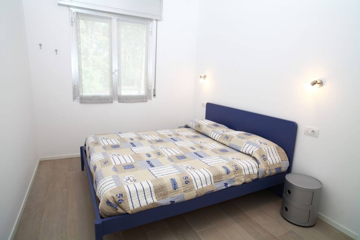 Un lit simple avec une tête de lit bleue, dans une pièce aux murs blancs, avec une fenêtre et des rideaux, et une petite poubelle grise.