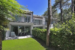 Et moderne hus med to etager, en altan, græsplæne og frodig beplantning omkring.