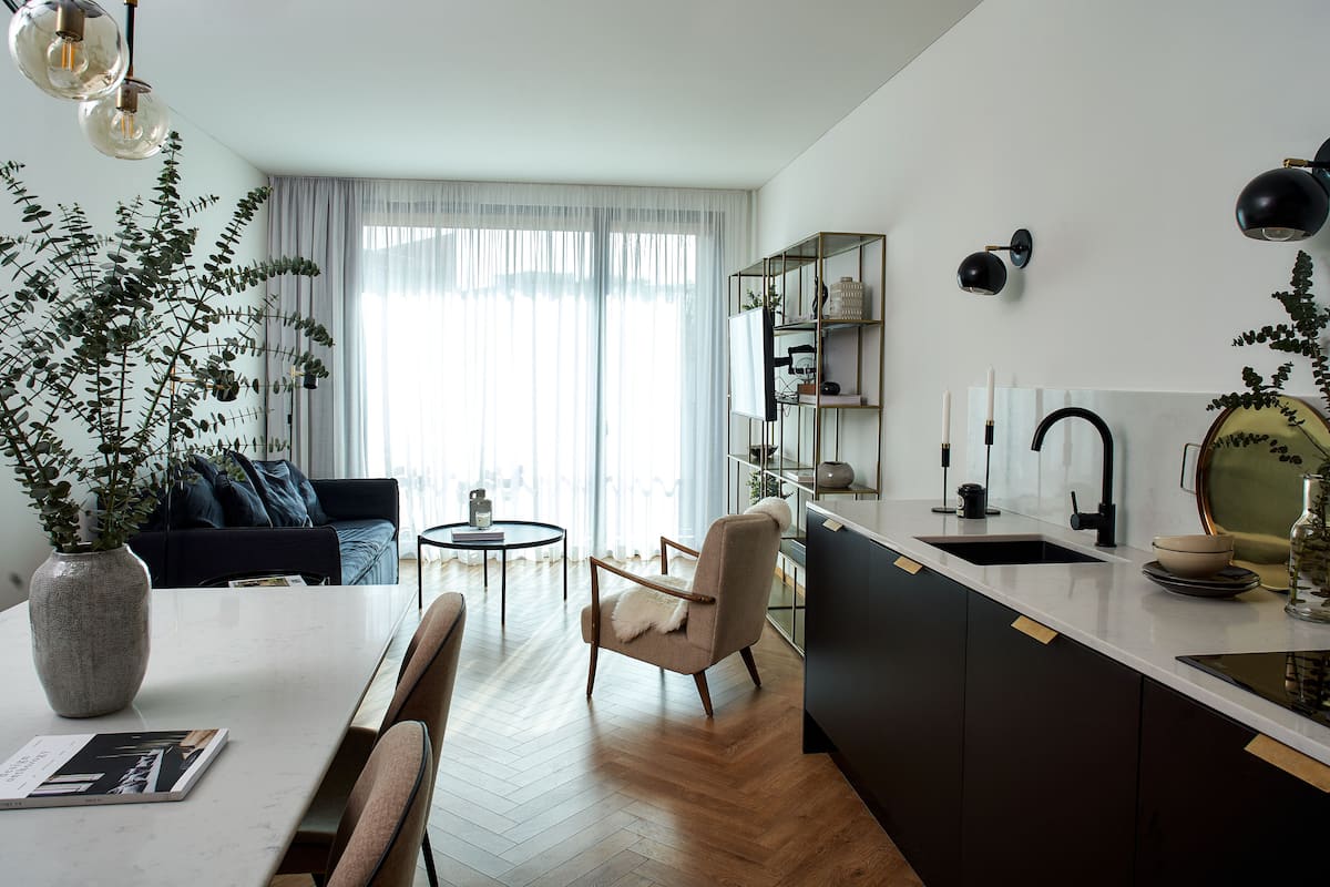 A modern kitchen with a white countertop, a sink, and a dining area with a table and chairs. There is a plant on the left and a bookshelf in the background.