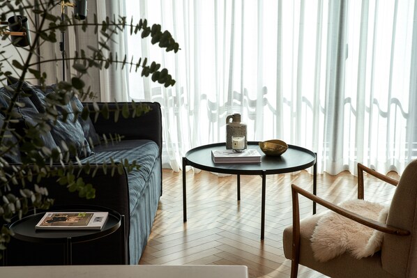 A modern living room with a sofa, a round coffee table, and a chair. There is a plant in the foreground, and a window with sheer curtains in the background.