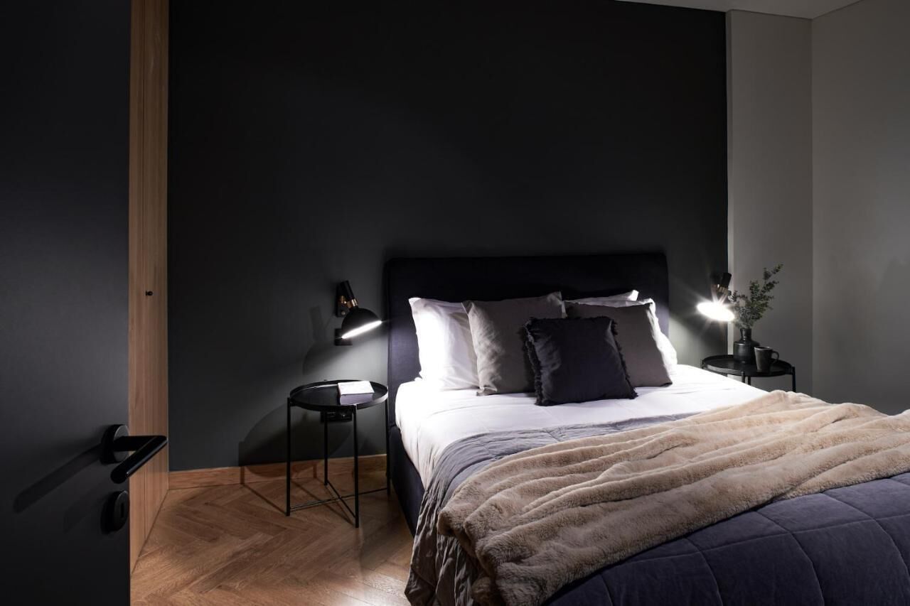A modern bedroom with a dark wall, a wooden floor, a bed with a fluffy blanket, two bedside tables with lamps, and a small plant on one table.