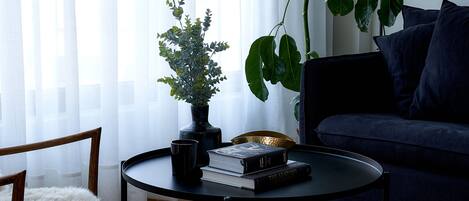 Un salón con un sofá azul oscuro, una mesita de centro con libros y una planta pequeña, un sillón blanco con un plaid suave y una planta verde grande.