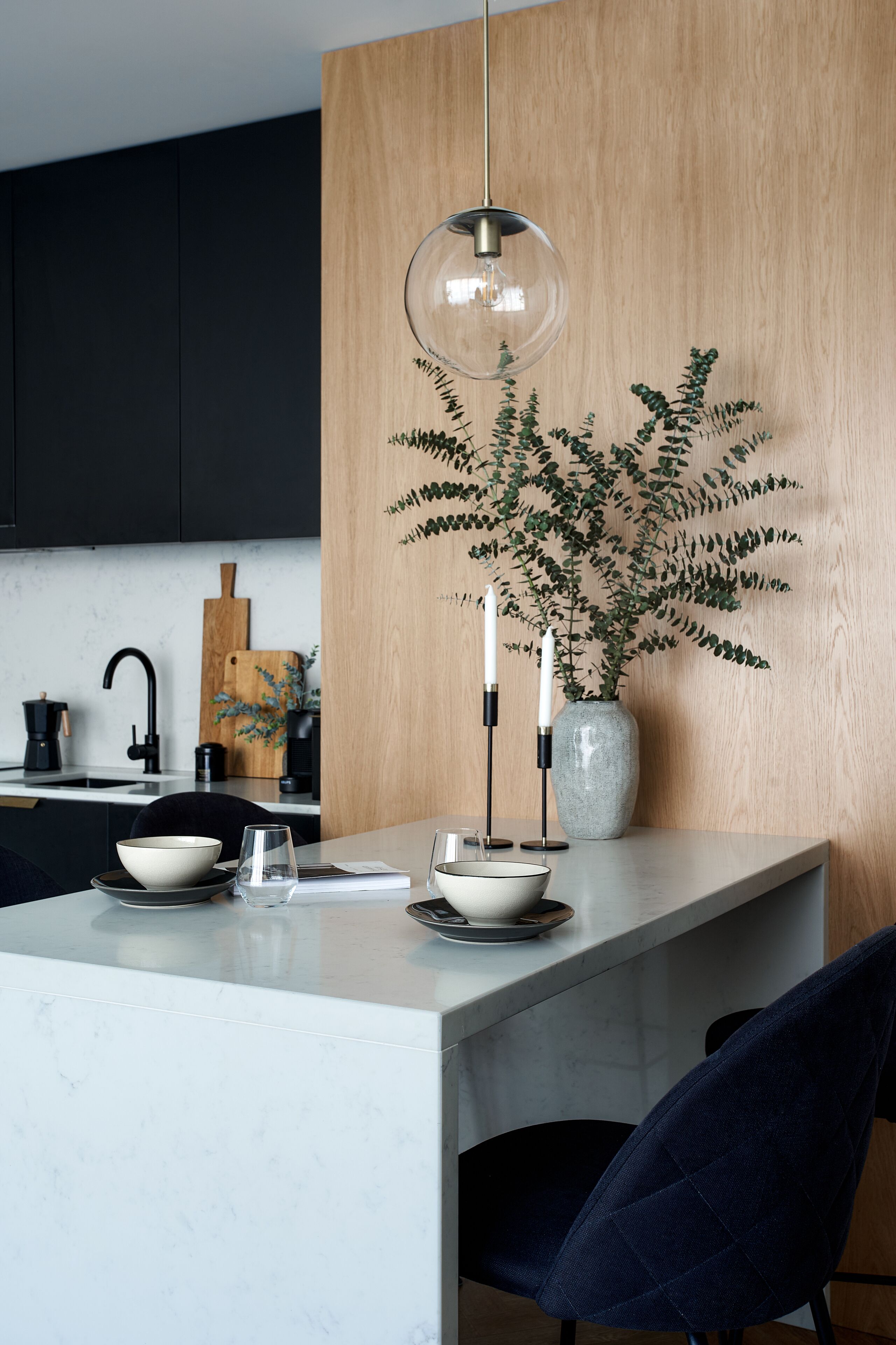 A modern kitchen with a central island, black cabinets, a wooden accent wall, a hanging pendant light, a vase with greenery, and a coffee maker.