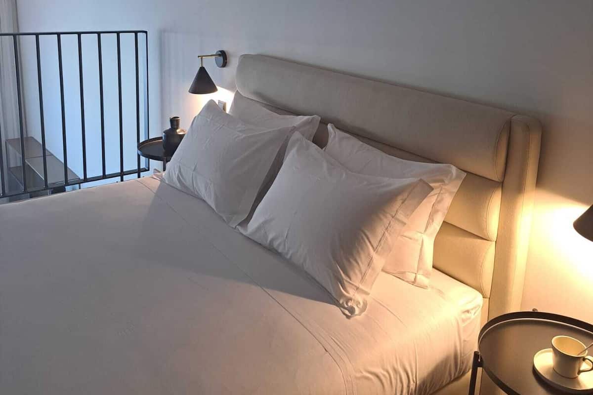 A neatly made bed with white linens, a bedside table with a lamp, and a small round table with a cup.