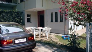 A parked car in front of a building with a patio area, a small table and chairs, and a flowering plant. - Villa Isolina (Lignano Sabbiadoro)