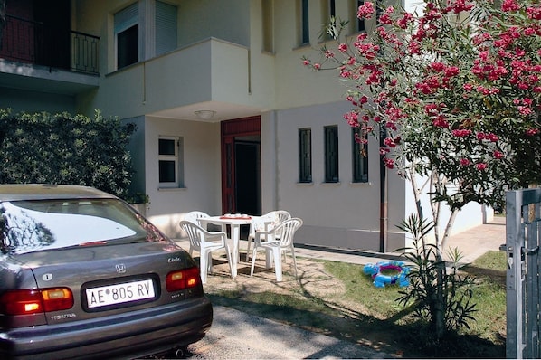 A parked car in front of a building with a patio area, a small table and chairs, and a flowering plant.