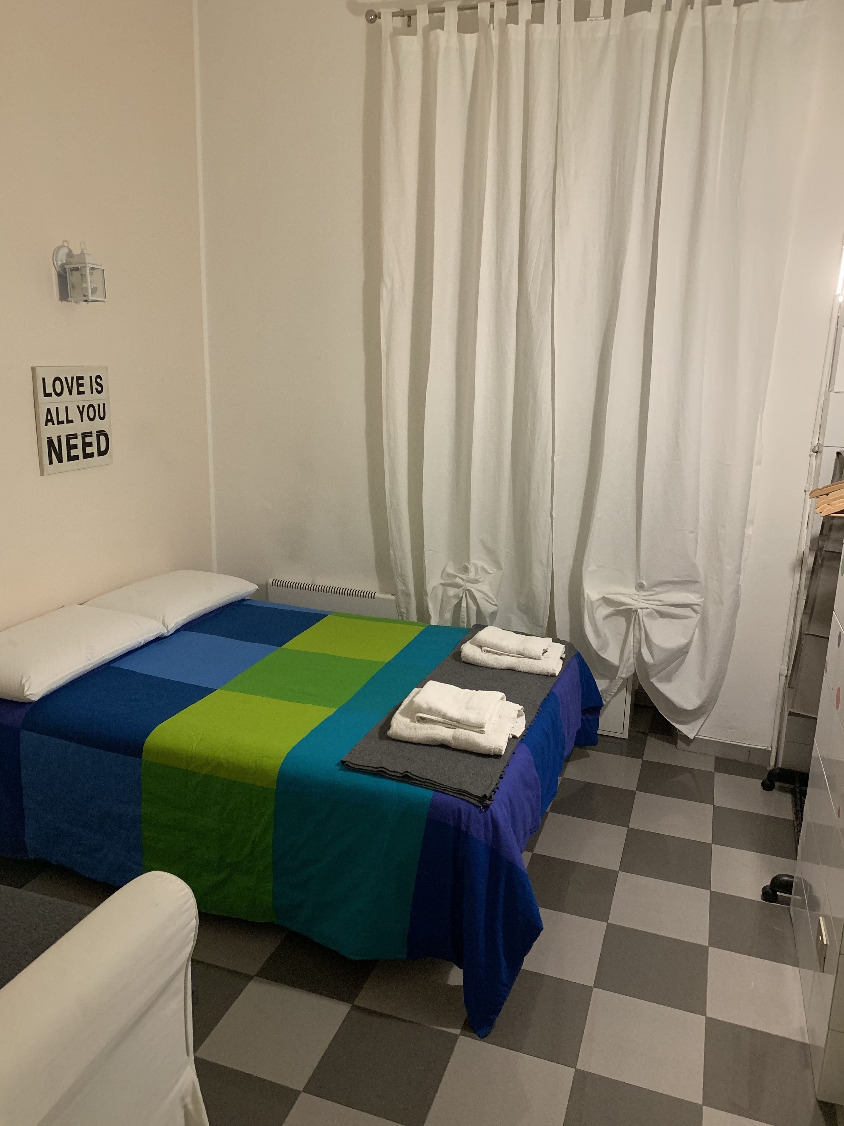 A hotel room with a checkered floor, a bed with a blue and green bedspread, white towels, and a sign on the wall that reads "LOVE IS ALL YOU NEED".