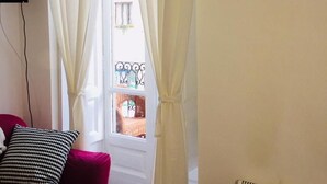 A room with a red sofa, white curtains, a window with a view of buildings, and a wall-mounted light fixture. - Bed2Go (Turin)